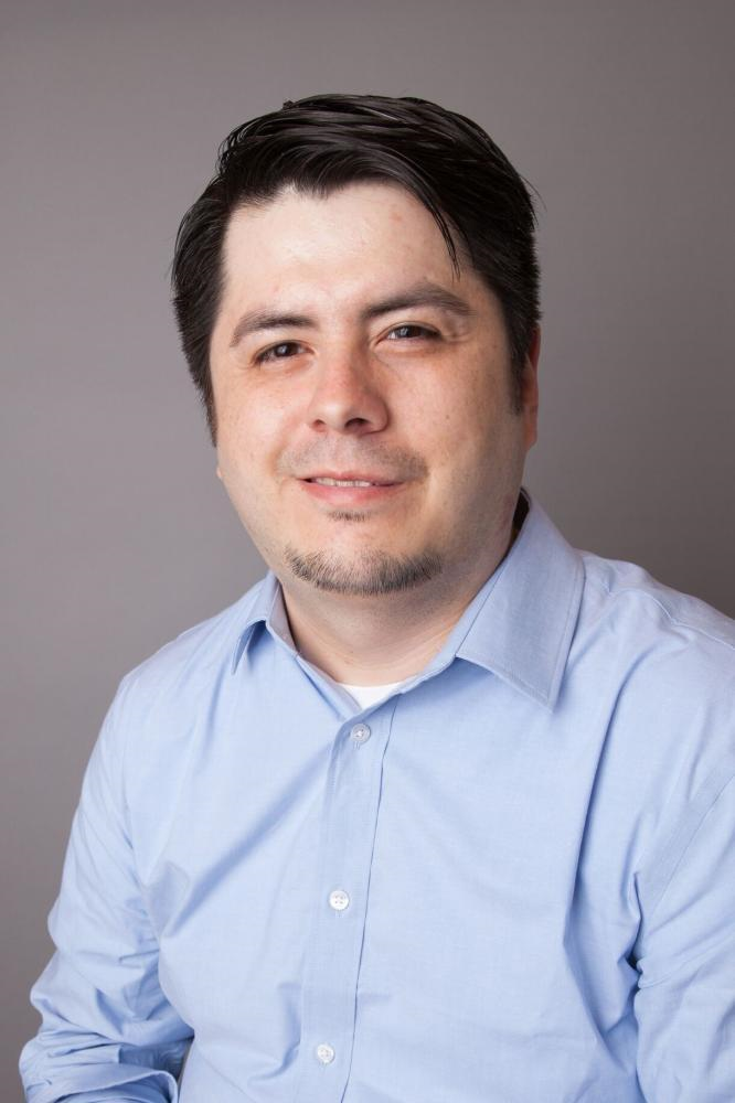 Formal portrait of Alisandro wearing a blue dress shirt with open collar. He has dark hair worn in a side part and a short facial hair along his jawline on his chin.