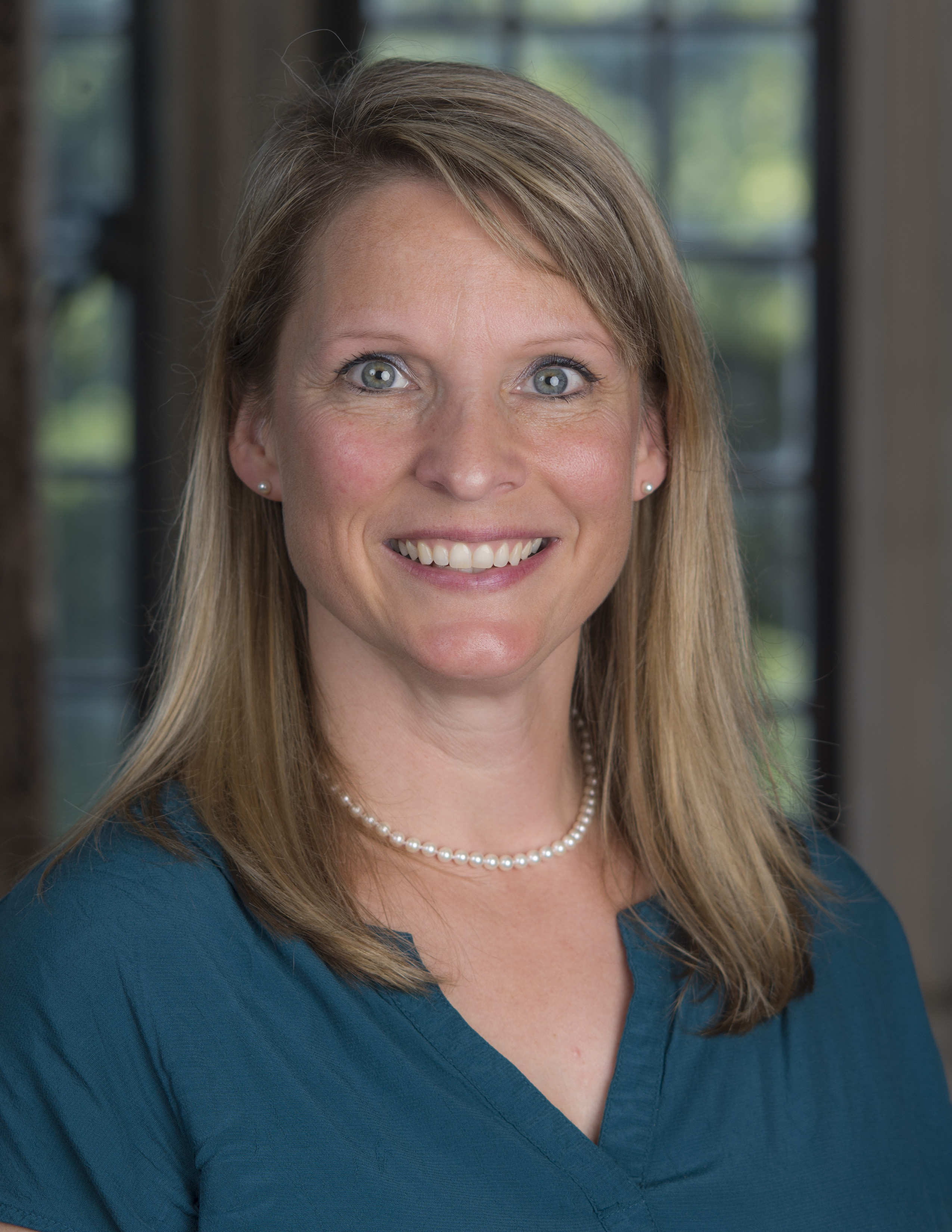 Smiling woman with shoulder length dark blond hair. She is wearing a blue shirt and a pearl necklace with matching earrings.