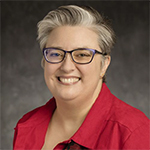 Smiling woman in a bright red shirt. Her hair is a mix of dark and light shades. Her hair is either short or pulled back. She is wearing glases with heavy dark frames.