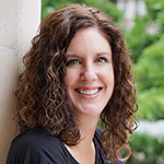 Smiling woman long curly brown hair. She is outdoors leaning against a wall.