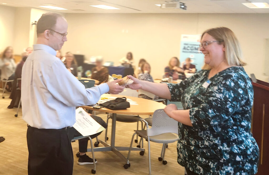 A man holding a certificate accepts a gift from a smiling woman. The woman is wearing glasses and a blue blouse with an abstract pattern. She has shoulder length blonde hair. The man has very short brown hair. He is wearing a light blue dress shirt, and black dress pants.  